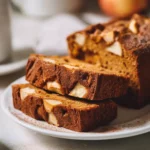 Moist pumpkin bread with apple chunks on a white plate, surrounded by cinnamon sticks and apples for a cozy fall vibe.