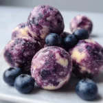 Blueberry Cheesecake Protein Bites 2 Blueberry cheesecake protein bites on a wooden board topped with fresh blueberries