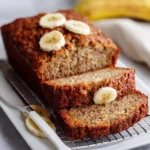 Slice of moist banana bread on a wooden table with ripe bananas in the background